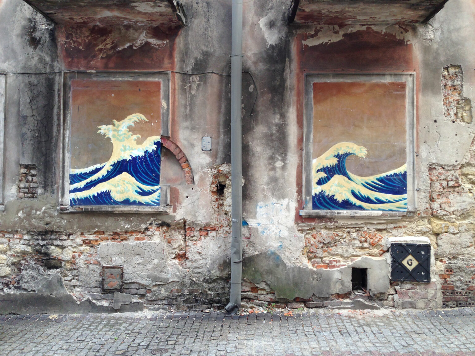 Auf die verbarrikadierten Fenster eines grauen Hauses, hat jemand leuchtend blau das Meer gemalt. Es ist als ob im Haus die Wellen wogen.